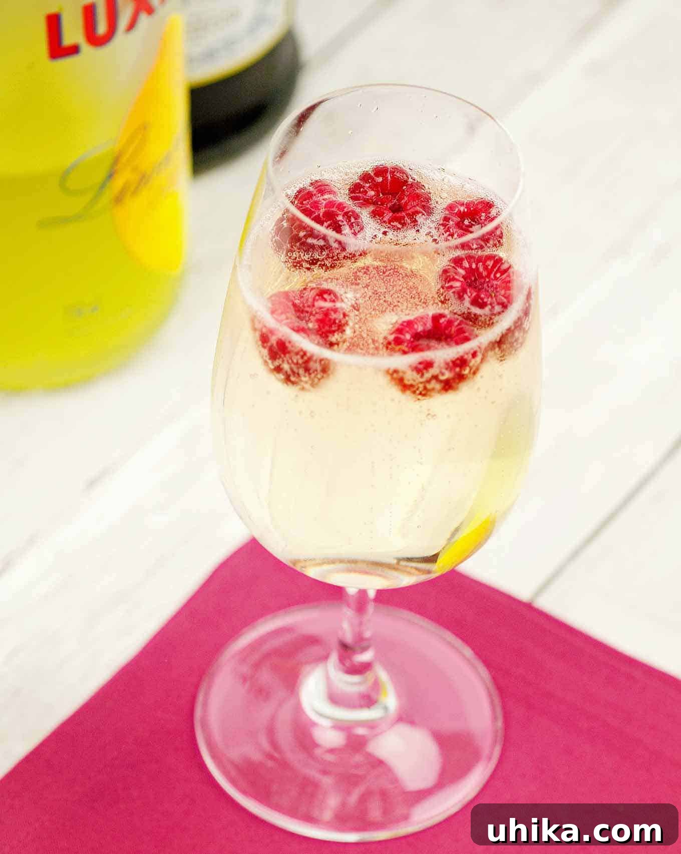 Fruity Prosecco Sparklers 2 A champagne flute filled with a sparkling cocktail of prosecco and limoncello with fresh raspberries floating on top on a pink cocktail napkin, bottles in the background.