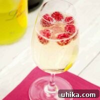 A champagne flute filled with a sparkling cocktail of prosecco and limoncello with fresh raspberries floating on top on a pink cocktail napkin, bottles in the background.