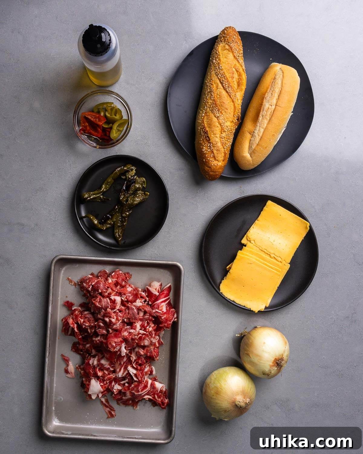 Effortless Philly Cheesesteak 4 A flat lay photograph displaying all ingredients required for a Philly Cheesesteak recipe: cooking oil, a jar of pickled cherry peppers, fresh Italian long hot peppers, soft hoagie rolls, thinly sliced raw ribeye steak, slices of American cheese, and chopped yellow onions.