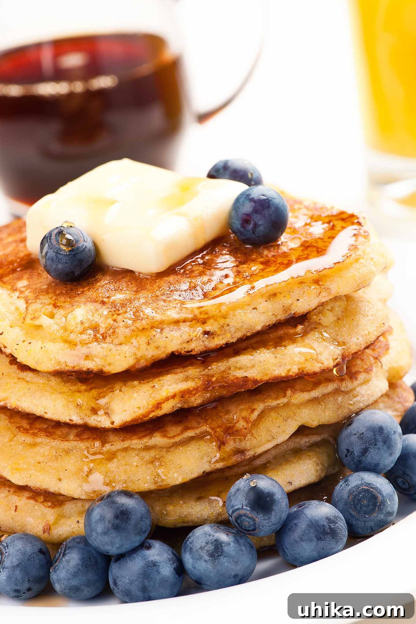 Golden Cornmeal Flapjacks 2 A stack of golden cornmeal pancakes on a white plate, generously topped with a melting pat of butter and rich maple syrup, garnished with fresh, vibrant blueberries. The pancakes feature a delicate crisp exterior and fluffy interior, promising a delightful breakfast.