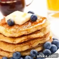 A stack of cornmeal pancakes on a plate, topped with fresh blueberries and a pat of butter, and drizzled with maple syrup, a small pitcher of syrup and a glass of orange juice in the background.