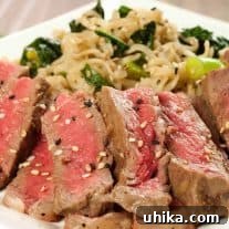 Sliced mirin-marinated strip steak topped with sesame seeds, served alongside scallion and kale ramen noodles on a white plate.