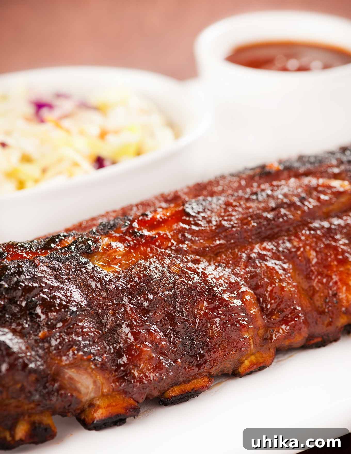 Fall-Off-The-Bone Oven Ribs 2 A close side view of a whole rack of baby back ribs, oven baked, glazed with homemade bourbon bbq sauce, coleslaw and more bbq sauce in the background.