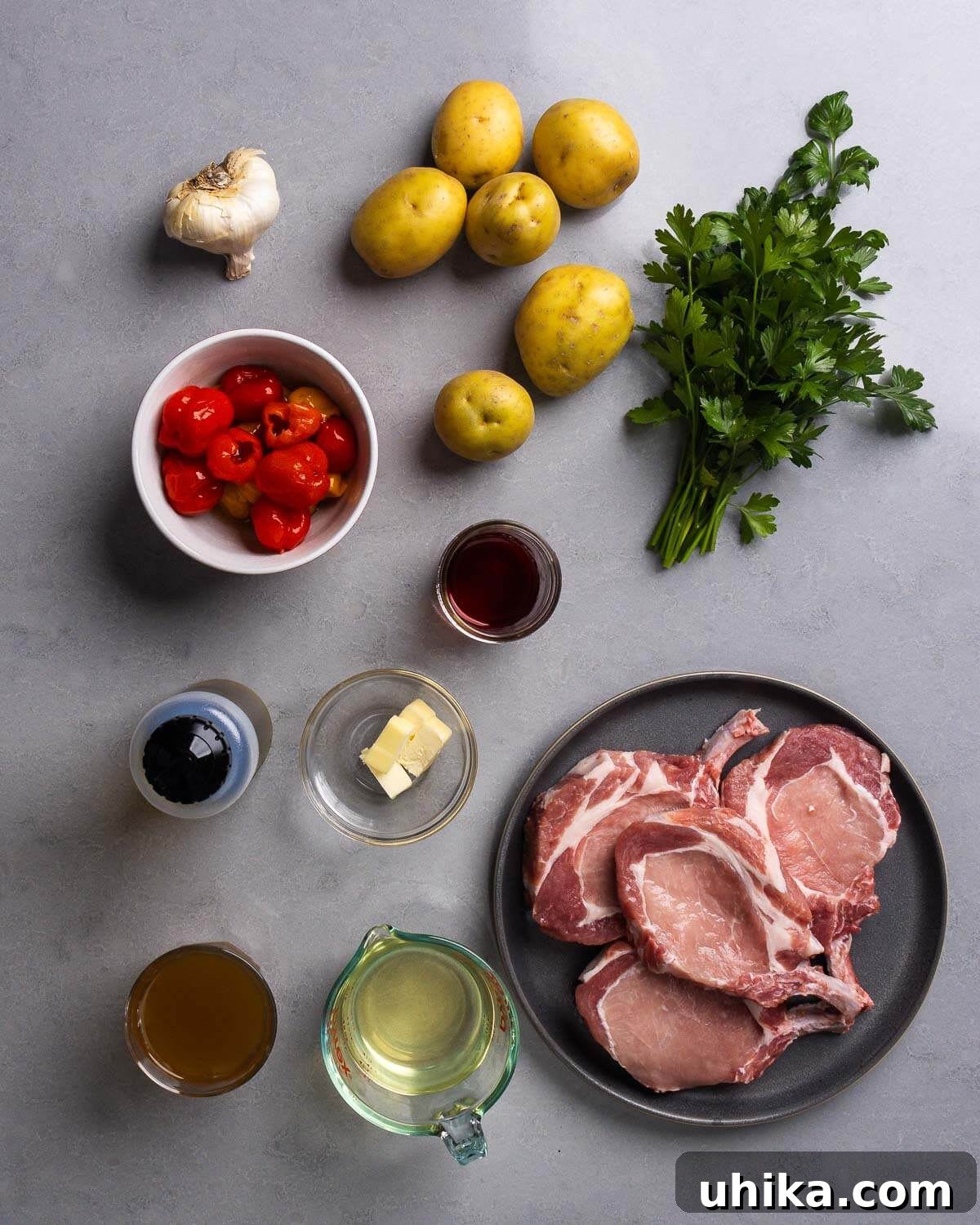 Sweet Heat Peppadew Pork with Crispy Roasted Potatoes 4 A flat lay photograph displaying all the fresh ingredients needed for the recipe: garlic cloves, quartered potatoes, fresh parsley, jarred peppadew peppers, red wine vinegar, olive oil, butter, a bottle of white wine, chicken stock, and raw bone-in pork chops.