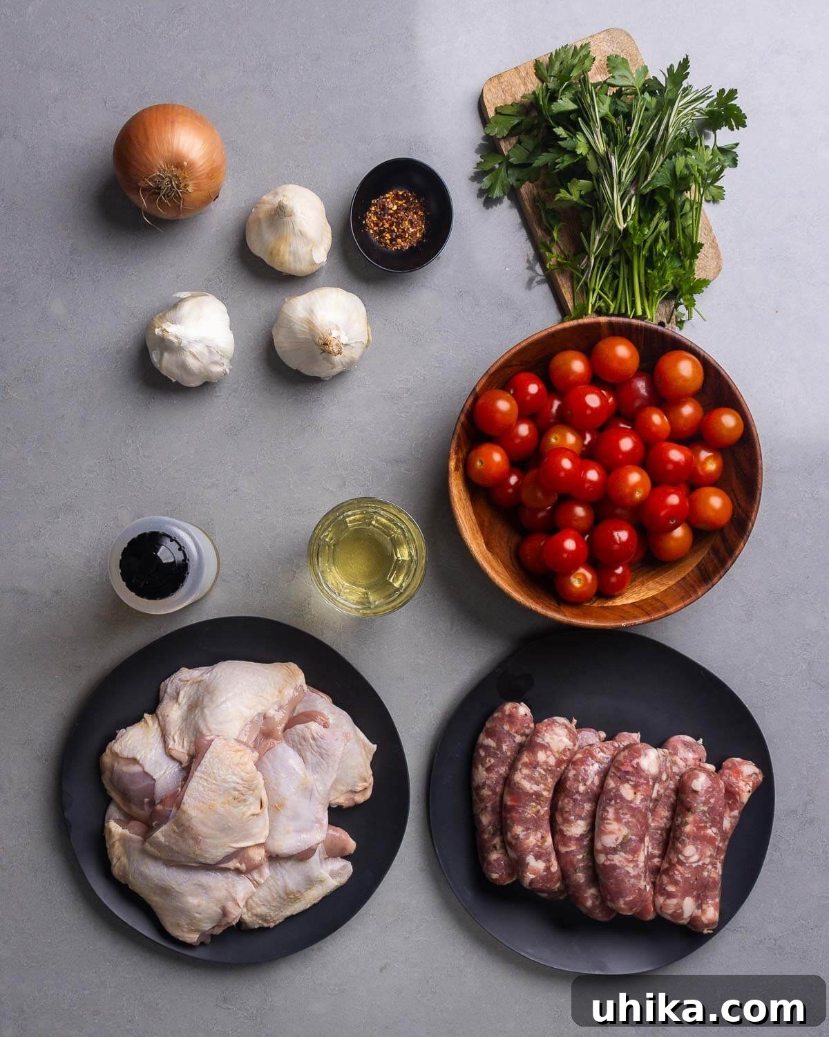 One-Pan Rosemary Chicken with Sausage and Burst Tomatoes 4 A flat lay of all ingredients for Rosemary Chicken with Sausage and Tomatoes: onion, garlic, hot red pepper flakes, parsley, rosemary, cherry tomatoes, olive oil, white wine, chicken thighs, and Italian sausage.