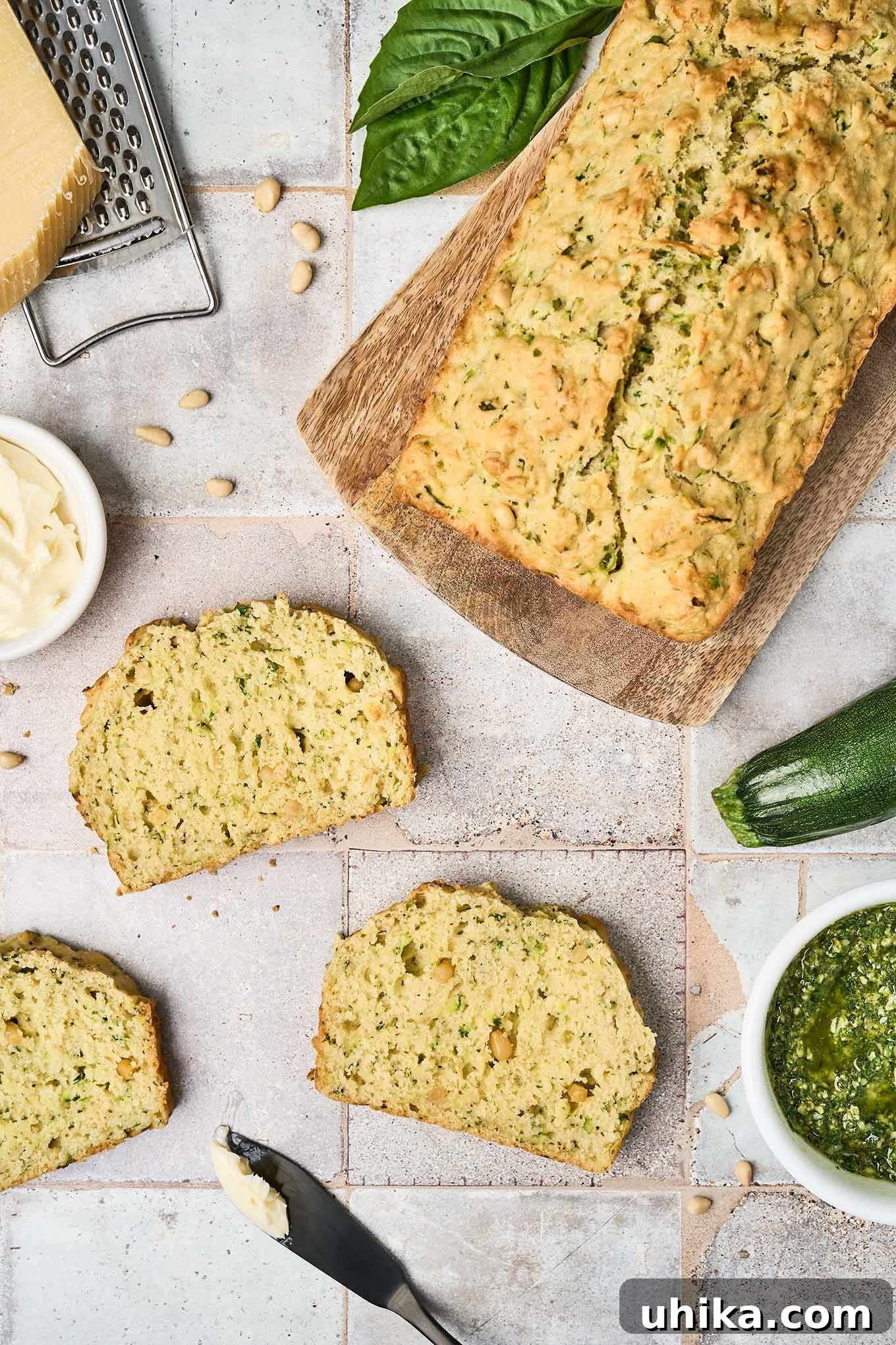 Hearty Savory Zucchini Bread 2 Three slices of savory pesto zucchini bread on a tile surface surrounded by a cutting board with the remainder of the loaf on it, a whole zucchini, a bowl of pesto, a dish of butter, Parmesan and a grater, pine nuts and basil leaves.