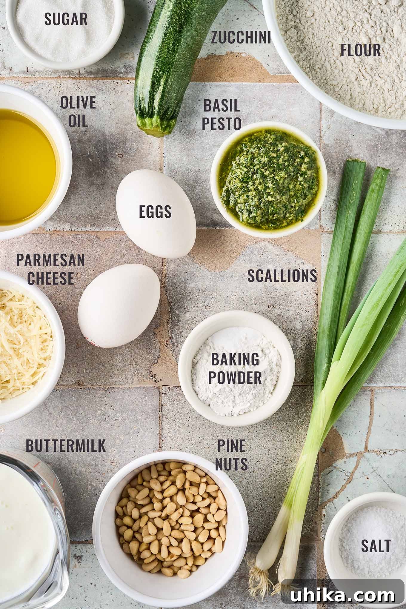 Hearty Savory Zucchini Bread 3 A labeled, overhead photo of the ingredients needed for savory pesto zucchini bread; flour, eggs, baking powder, basil pesto, scallions, pine nuts, buttermilk, Parmesan cheese, and olive oil.