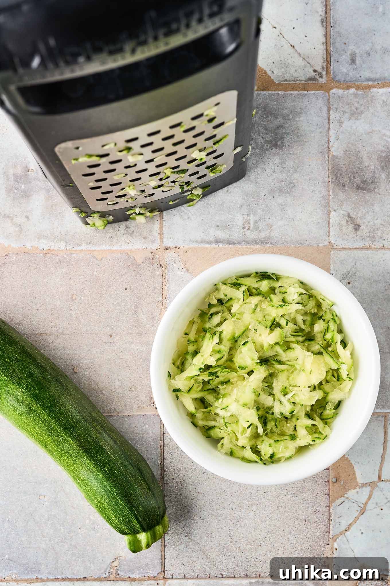 Hearty Savory Zucchini Bread 4 Coarsely grated zucchini in a bowl next to a box grater showing the large holes, a whole zucchini to the side.