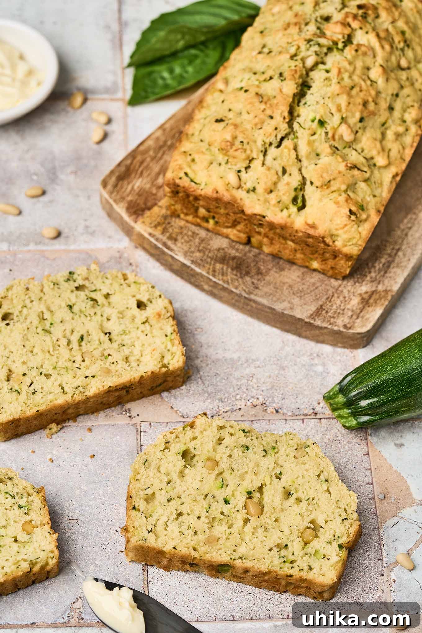 Hearty Savory Zucchini Bread 8 A close view of slices of savory zucchini bread on a tile countertop, a knife with butter on it, and a cutting board with a loaf of zucchini bread on it.