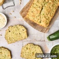 Three slices of savory pesto zucchini bread on a tile surface surrounded by a cutting board with the remainder of the loaf on it, a whole zucchini, a bowl of pesto, a dish of butter, Parmesan and a grater, pine nuts and basil leaves.