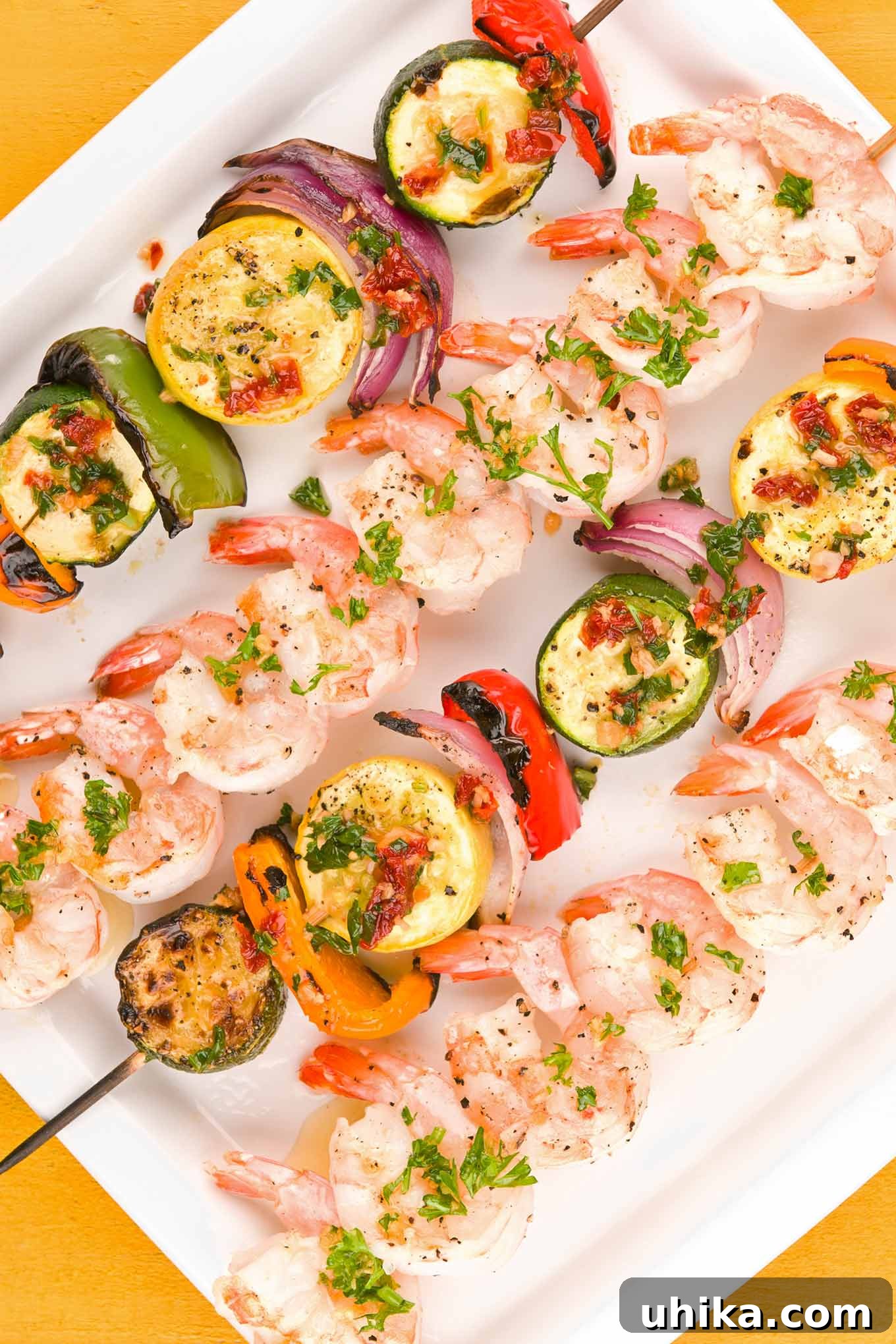 Skewers of grilled shrimp and vibrant vegetables, including zucchini, yellow squash, bell pepper, and red onion, all brushed with a savory garlic butter and arranged on a square white serving plate.