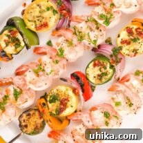 Skewers of grilled shrimp and vegetables (zucchini, yellow squash, bell pepper, and red onion) brushed with garlic butter arranged on a square white serving plate.