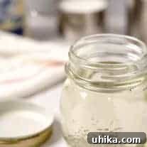 A jar of prepared simple syrup with the lid beside it, bag of sugar, measuring cup, and saucepan in the background