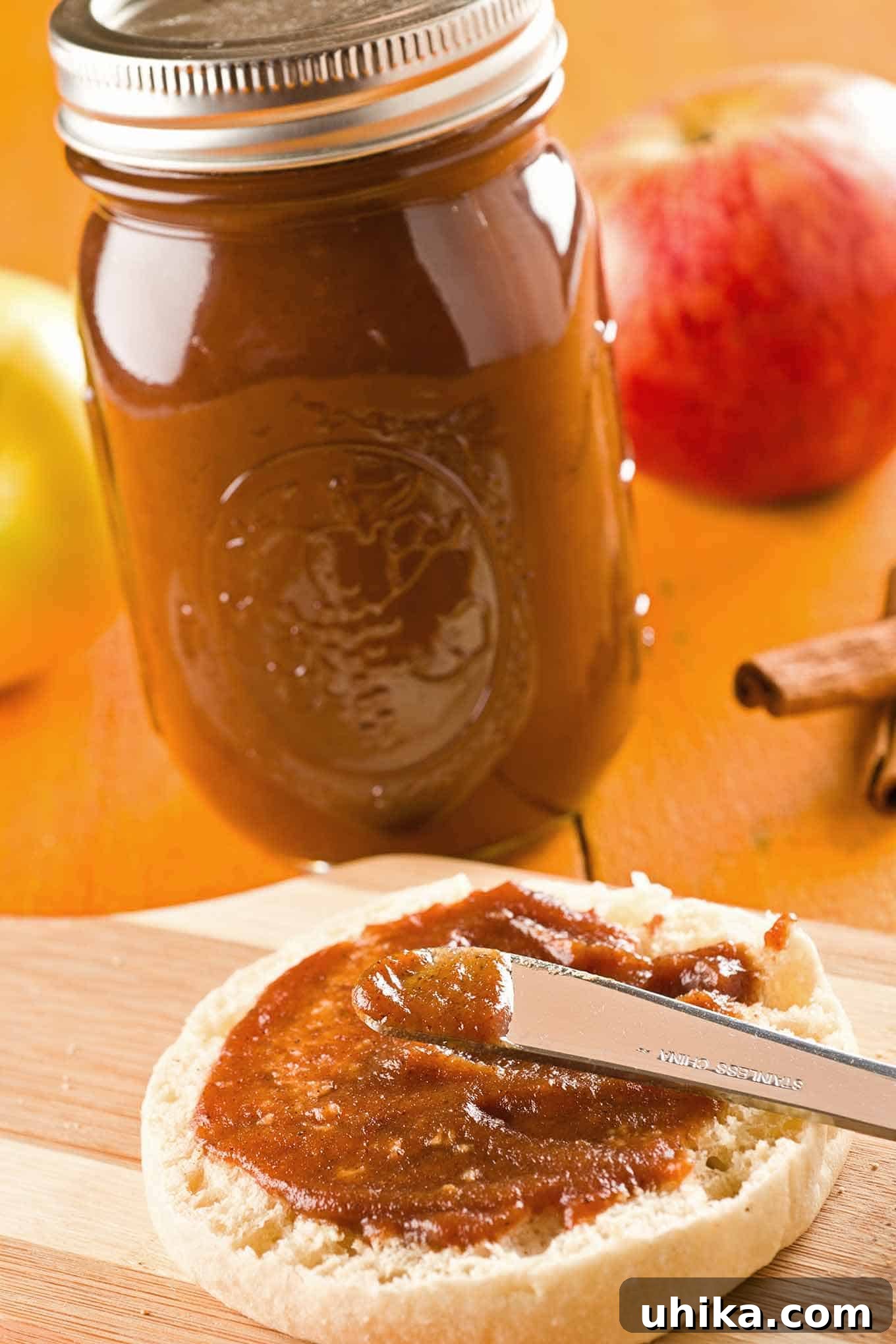Homemade Slow Cooker Apple Butter 2 Apple butter being spread on an English muffin with a jar of slow cooker apple butter in the background alongside fresh whole apples and cinnamon sticks.