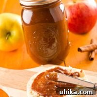 An English muffin on a cutting board being spread with homemade, slow cooker apple butter, a Mason jar filled with apple butter in the background, alongside whole apples and cinnamon sticks.