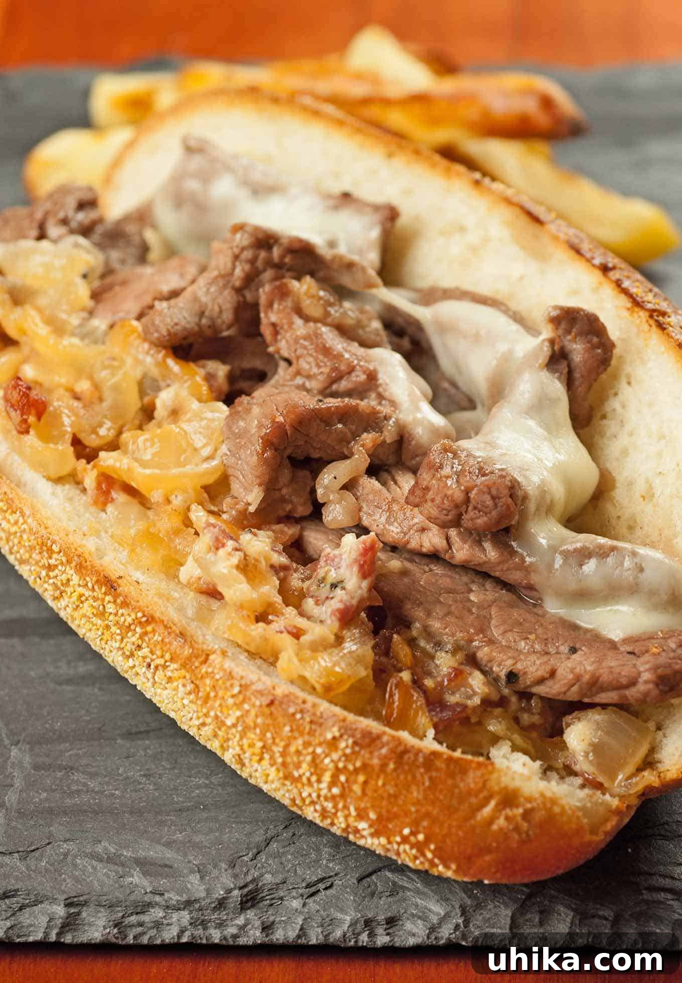 NY Strip Steak Sandwich with Zesty Bacon Horseradish Aioli 2 Strip steak sandwich with melted cheese, caramelized onions, and bacon-horseradish spread on a toasted hoagie roll, served with fries in the background.