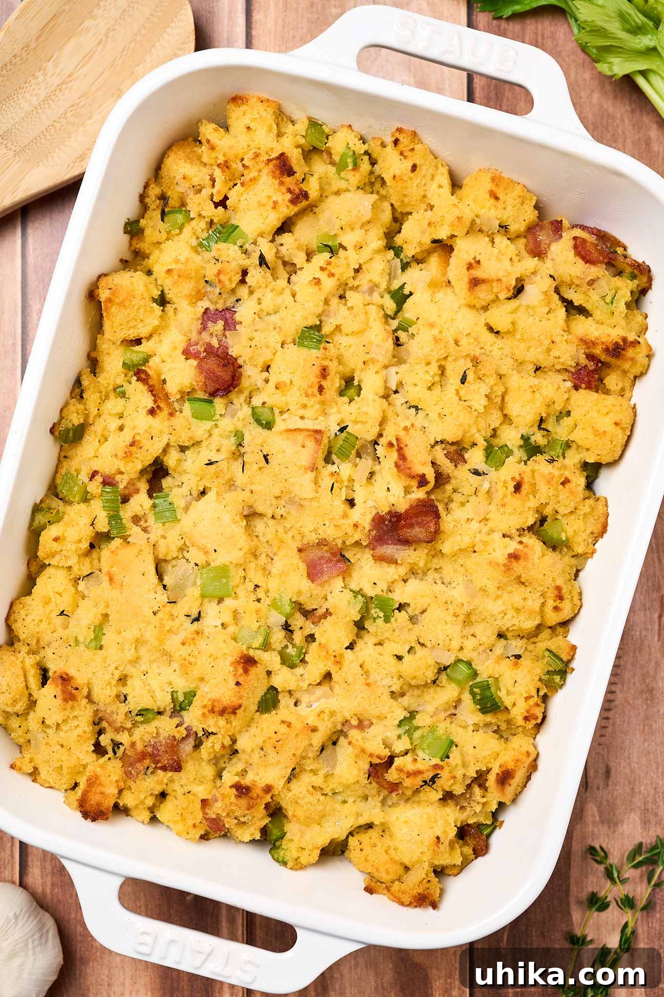 Smoked Bacon Cornbread Dressing 2 Homemade cornbread stuffing fresh from the oven in a white oblong casserole dish on a wooden surface surrounded by some ingredients including fresh thyme, celery, and garlic.