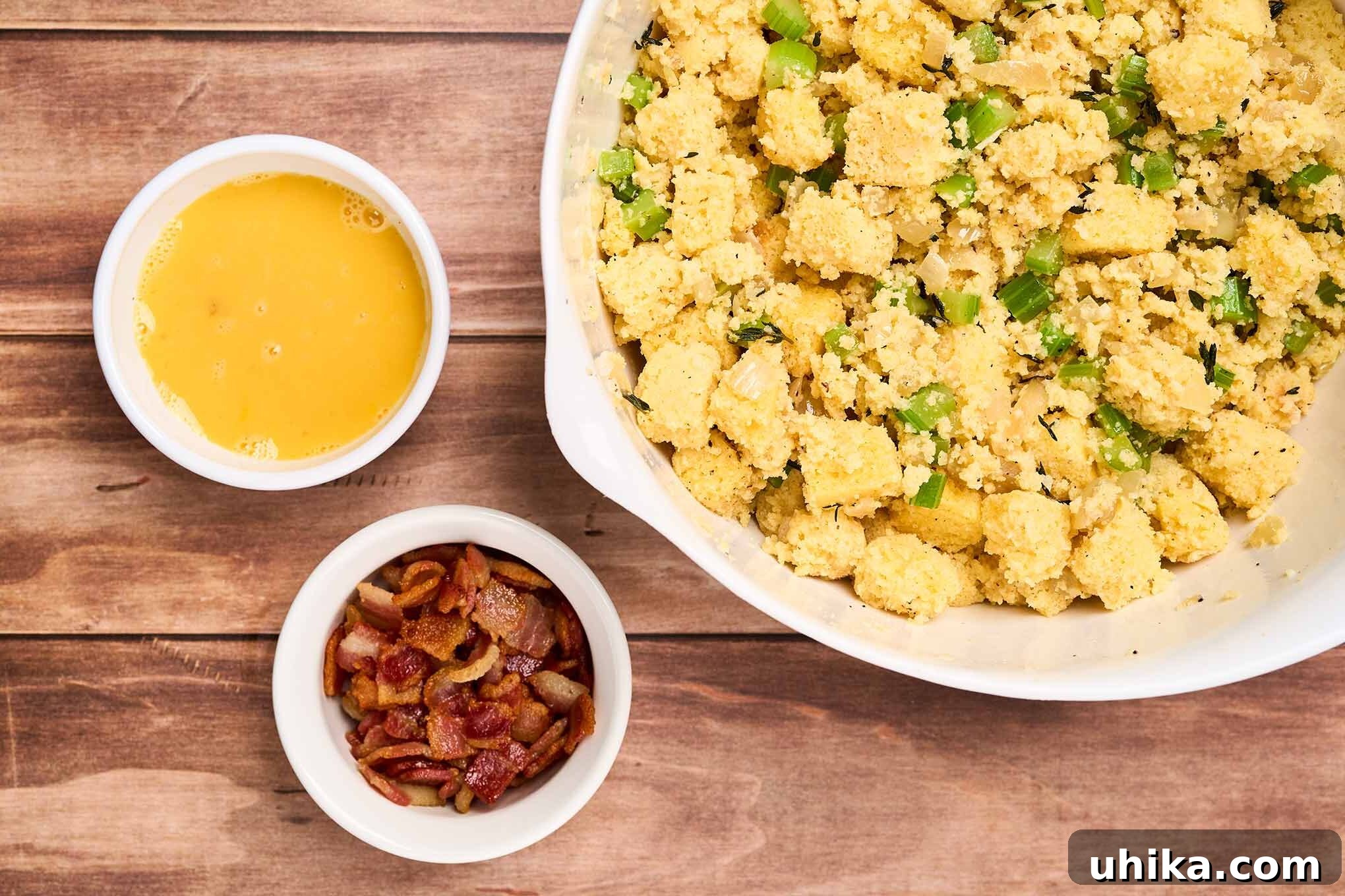 Smoked Bacon Cornbread Dressing 6 Cornbread stuffing preparation step three showing a bowl of moistened cubes of cornbread combined with aromatics alongside bowls of bacon bits and eggs.