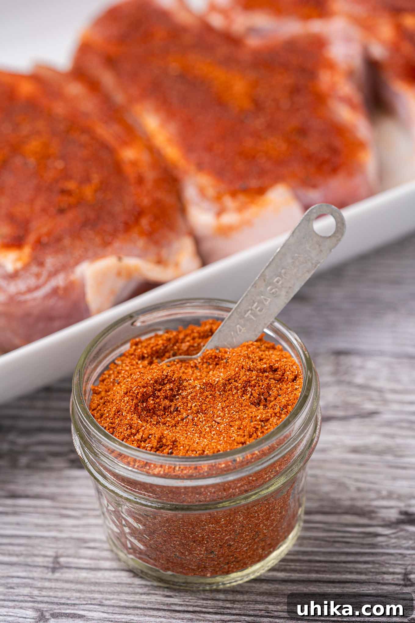 Effortless BBQ Flavor Blend 2 A glass jar of BBQ spice rub with a measuring spoon in it with rub-coated pork chops in the background.