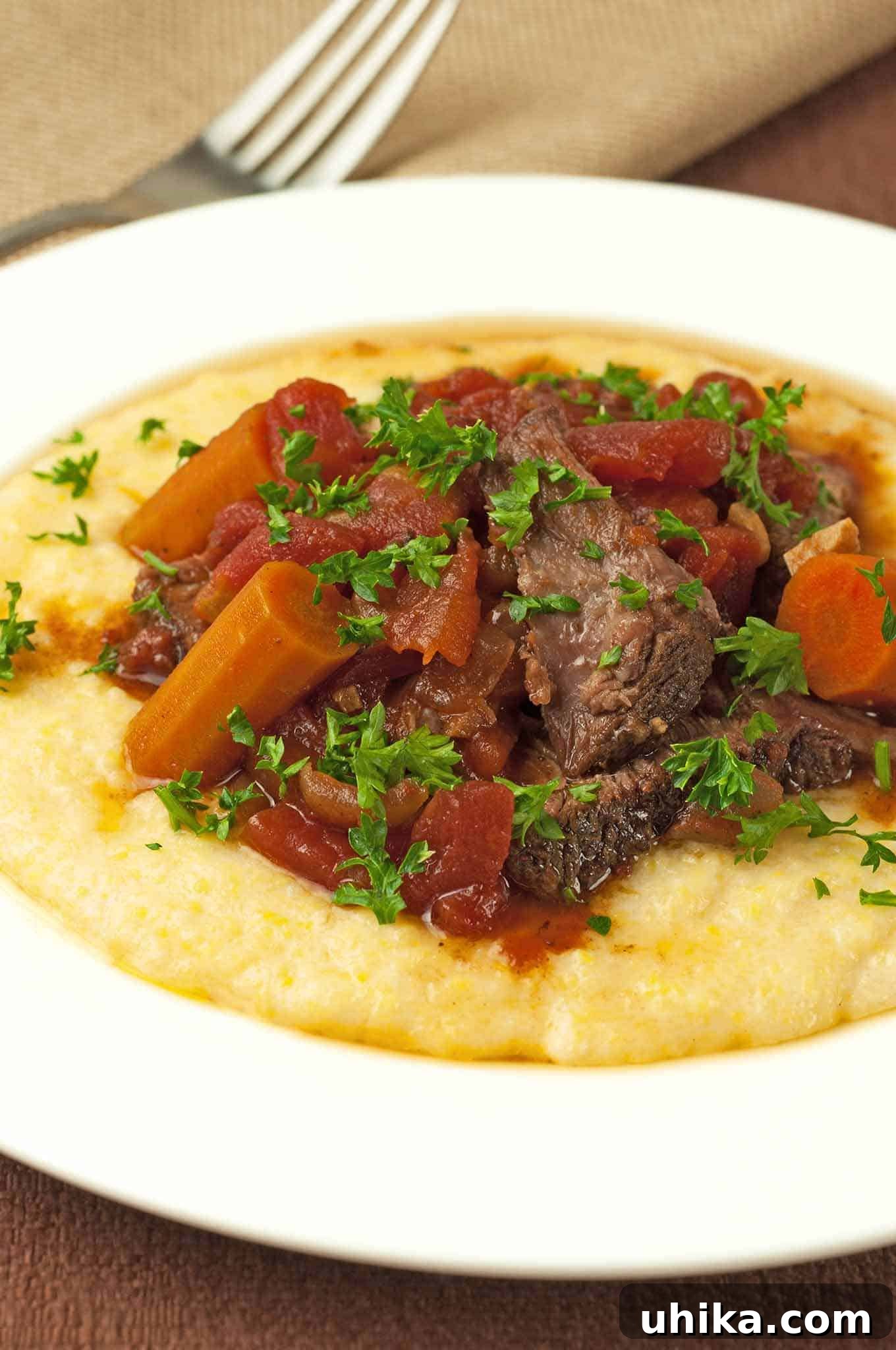 Italian pot roast served over creamy polenta, topped with tender carrots, stewed tomatoes, and chopped parsley on a white plate.