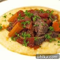 Italian pot roast served over creamy polenta, topped with tender carrots, stewed tomatoes, and chopped parsley on a white plate.