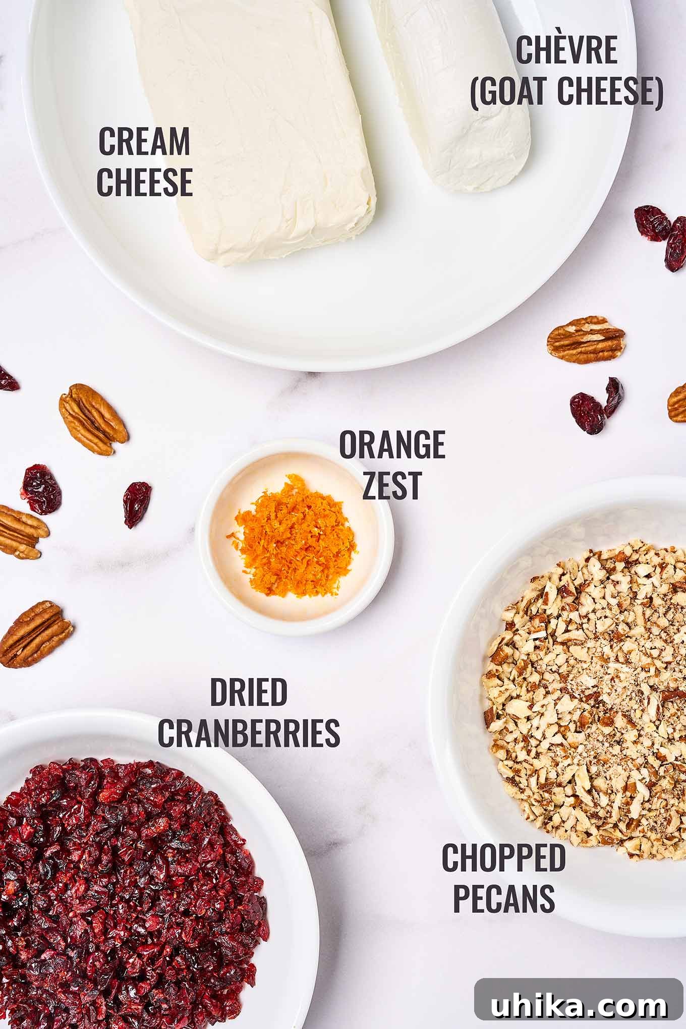 A labeled overhead view of the ingredients for a cranberry cheese ball including cream cheese, goat cheese, orange zest, dried cranberries, and pecans.