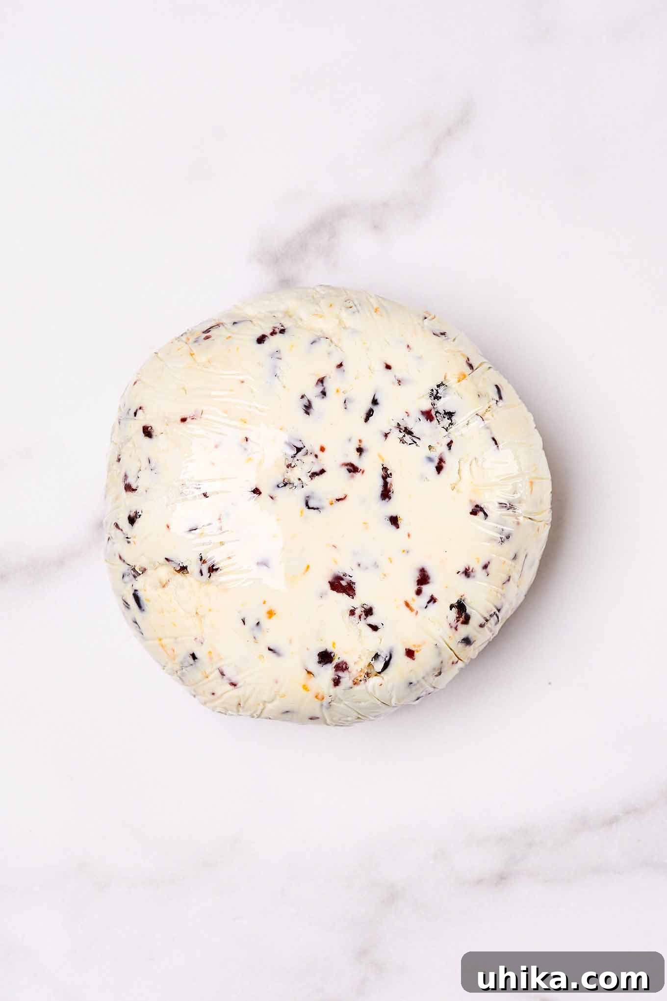 A cranberry cheese ball tightly wrapped in plastic wrap and ready to be refrigerated on a marble board.