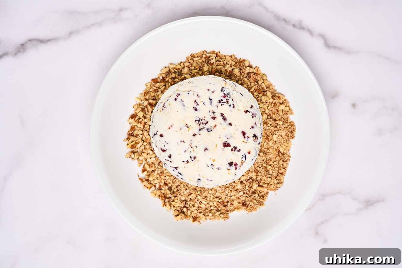 A cranberry cheese ball on a bed of chopped, toasted nuts on a plate, ready to be rolled.