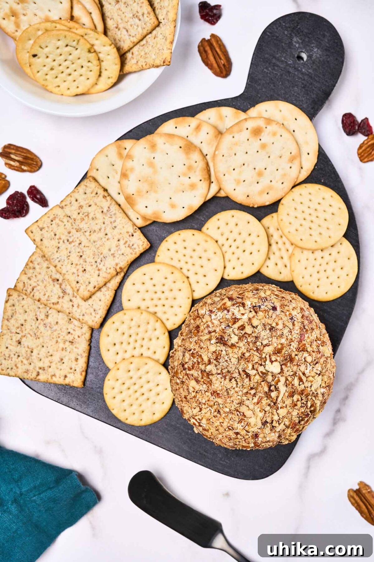 An overhead view of a cranberry cheese ball and crackers atop a black serving board. More crackers, toasted pecans, and a spreader surround the board.