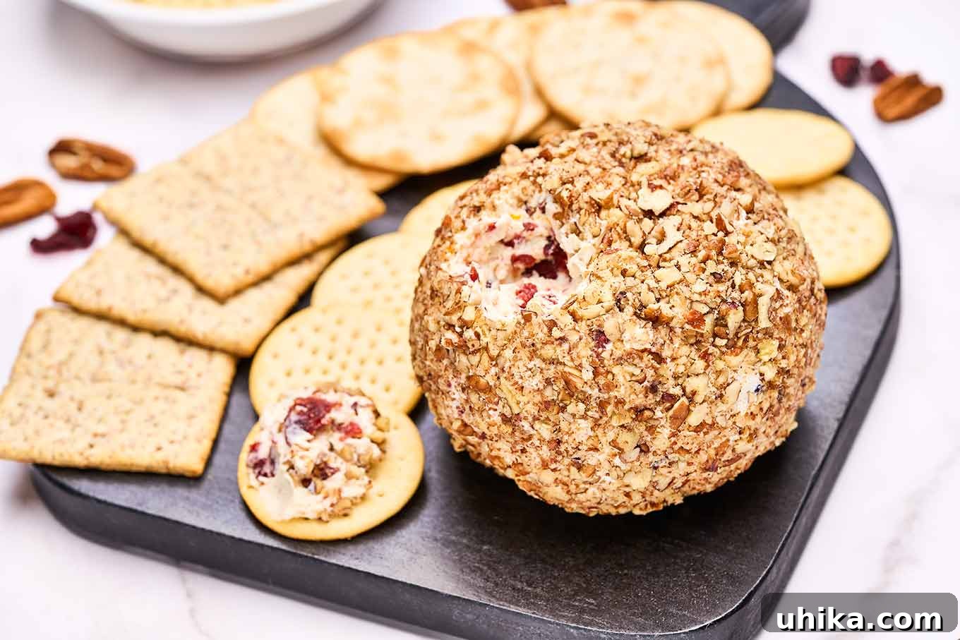A black serving board with a variety of crackers and a cranberry cheese ball rolled in toasted nuts. A small amount was removed and spread onto the cracker to the left of the cheese ball.