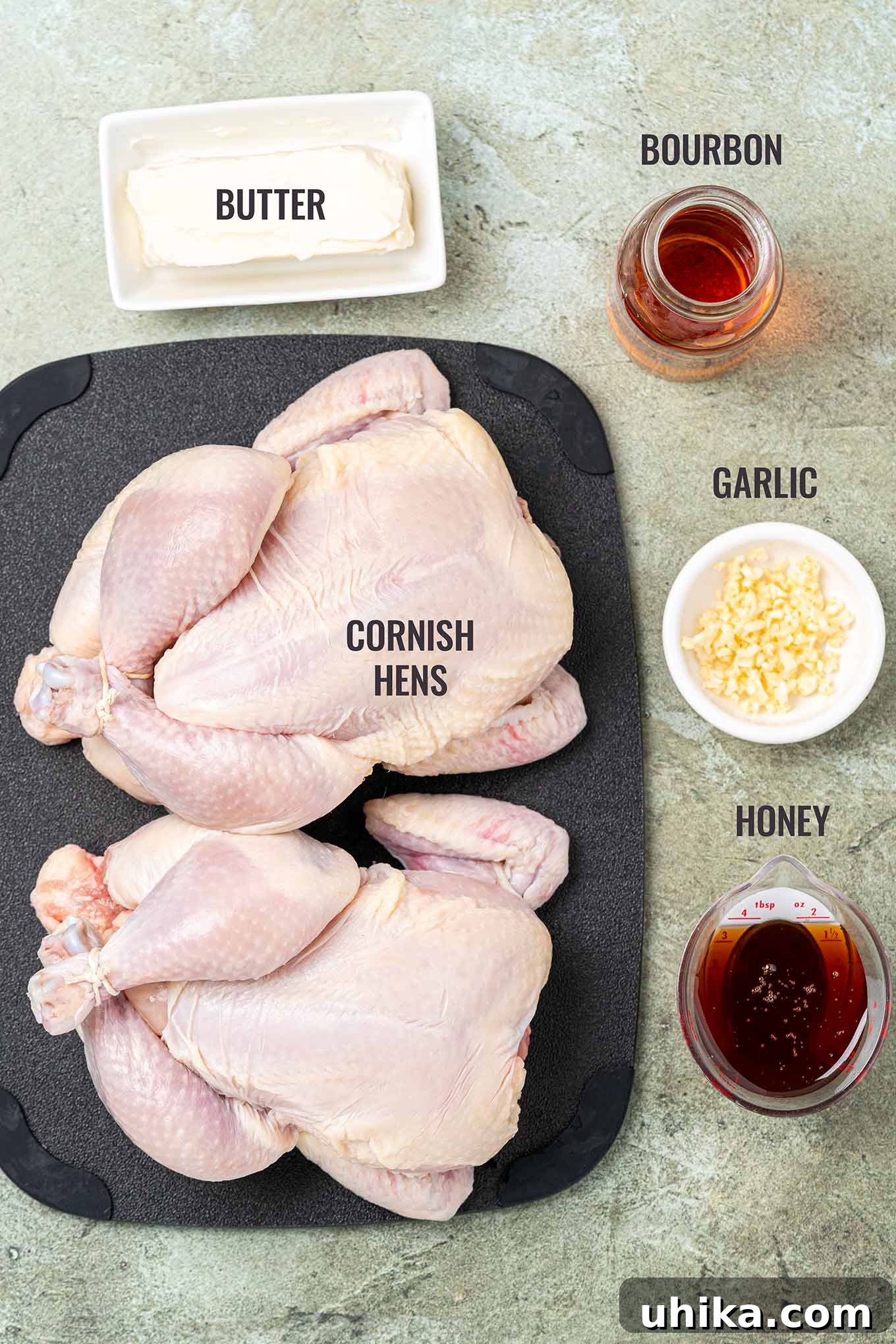 Golden Bourbon Glazed Cornish Hens 3 A labeled, overhead photo of the ingredients needed to make Cornish hens with bourbon glaze, including two cornish hens, butter, bourbon, garlic, and honey.