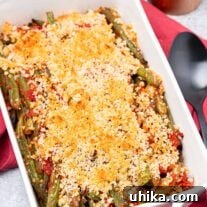 Fresh from the oven, a casserole of baked Italian-style green beans and tomatoes with crisp Parmesan-panko topping, a pepper mill and serving spoon in the background.