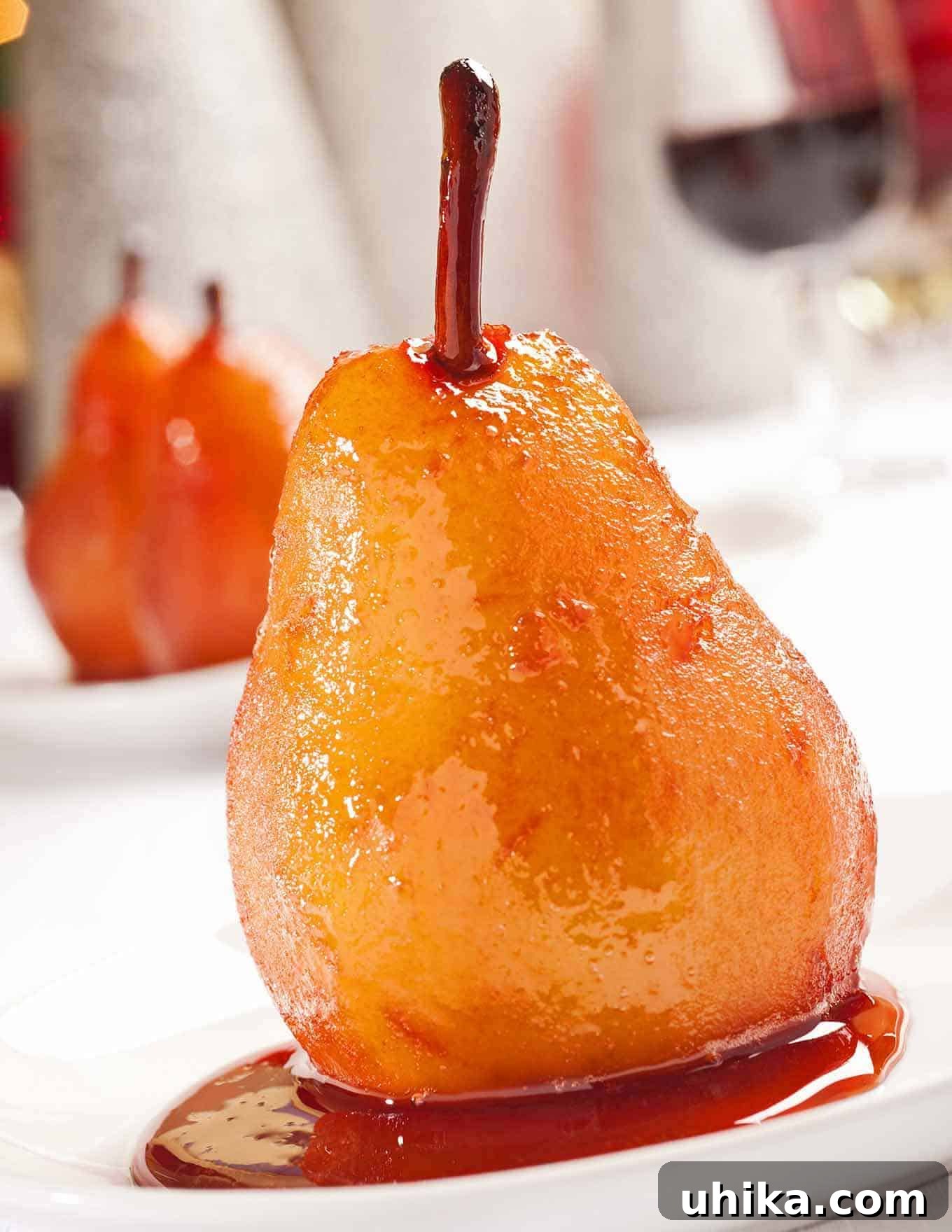 Ruby Port Pears 2 Port wine poached pear glistening in rich syrup on a white dessert plate, with more pears and red wine in the blurred background.