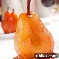 Port wine poached pear glistening in rich syrup on a white dessert plate, with more pears and red wine in the blurred background.