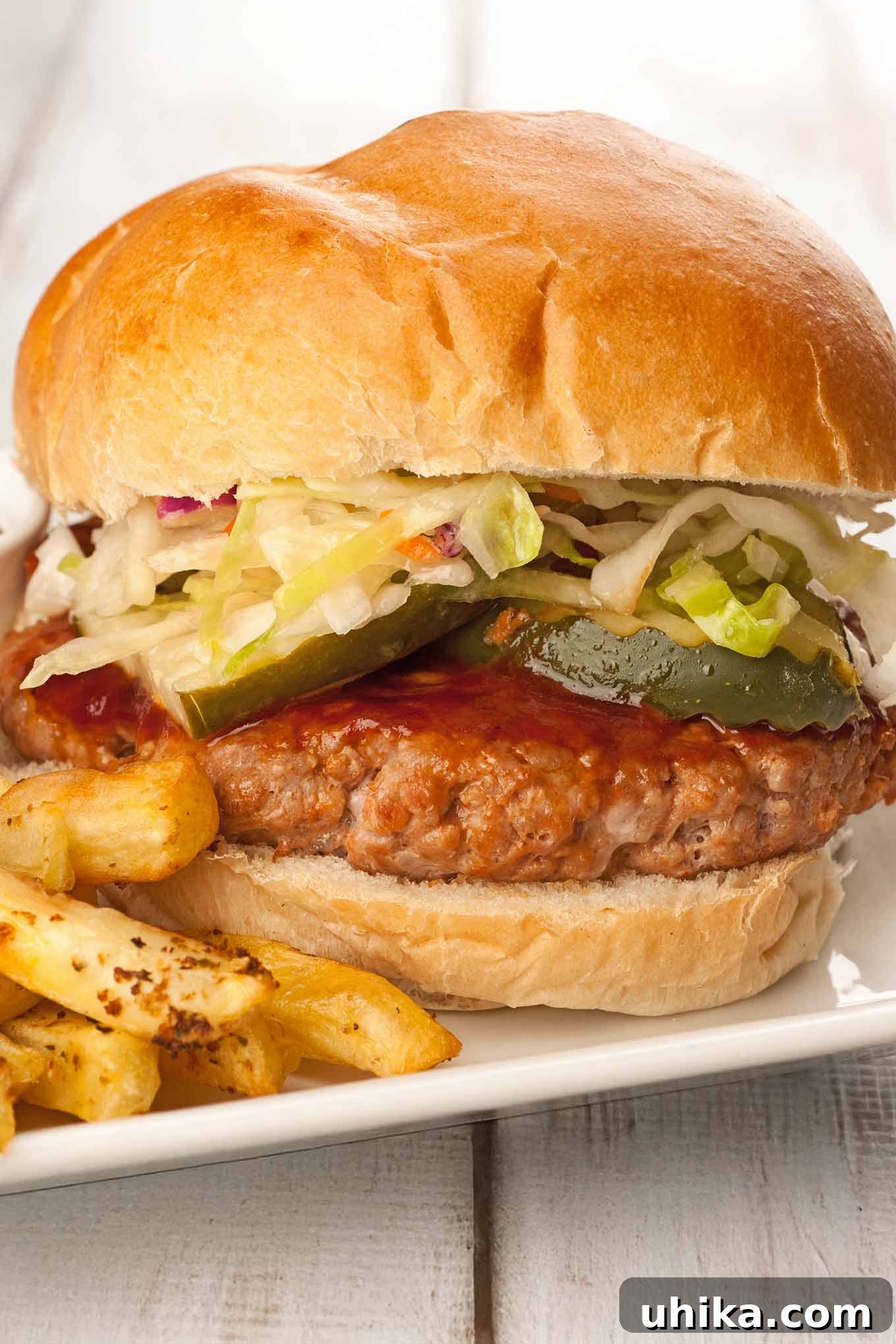 Flame-Kissed Pork Burgers 2 A close view of a grilled pork patty topped with bourbon bbq sauce, pickle slices, and coleslaw on a soft hamburger roll with fries on the side.