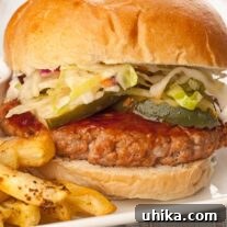 Flame-Kissed Pork Burgers 4 A close view of a grilled pork patty topped with bourbon bbq sauce, pickle slices, and coleslaw on a soft hamburger roll with fries on the side.