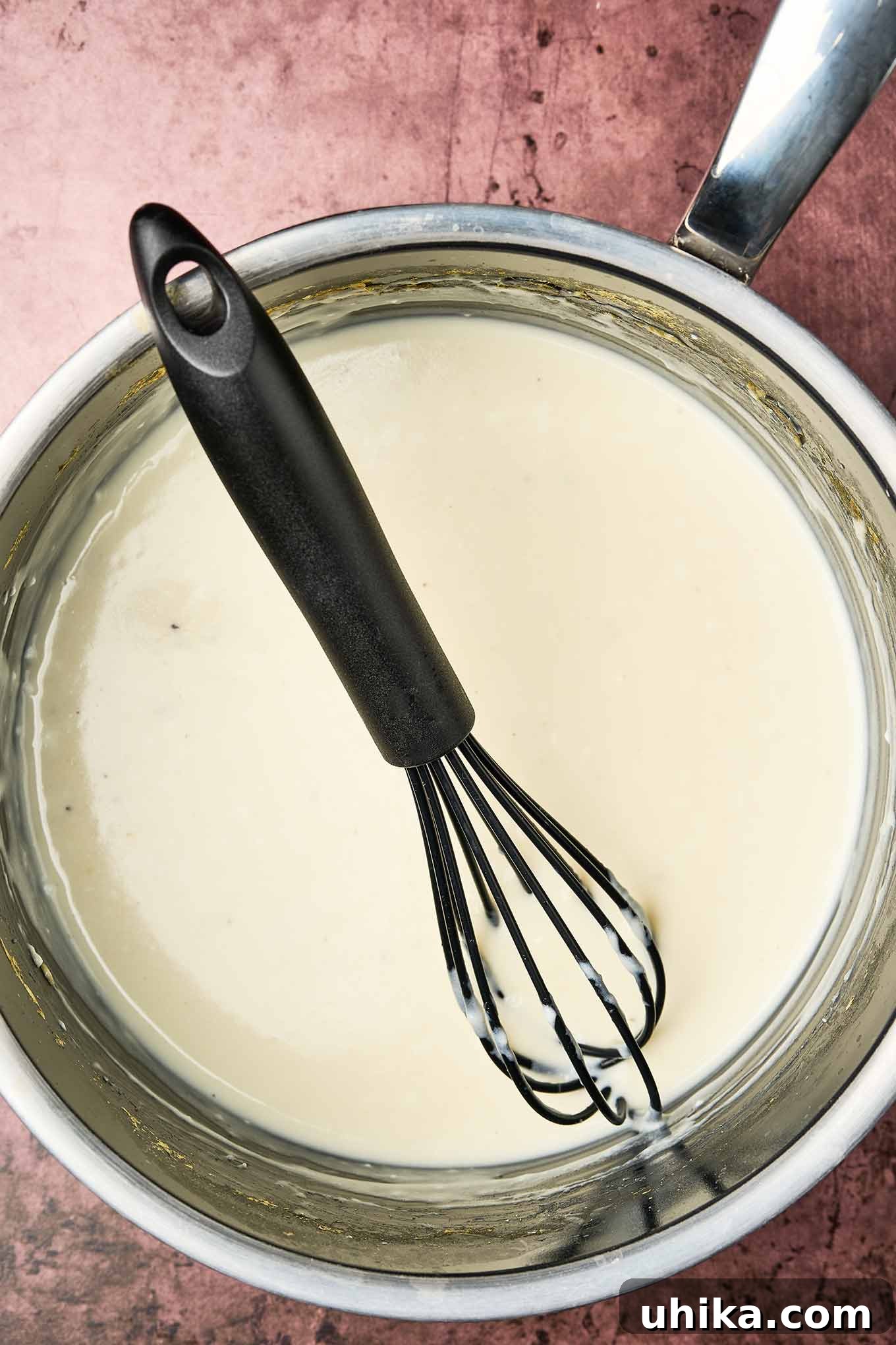Creamy Cottage Potato Gratin 4 Smooth and creamy white cheese sauce in a saucepan with a black whisk, shown from above, ready to be combined with potatoes and chives and baked.