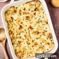 Golden-browned cottage cheese potato casserole in a white baking dish, topped with melted cheese and chopped chives, on a rustic surface with whole potatoes and a wooden spoon nearby.