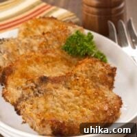 An oval plate with three breaded, oven-fried pork chops, garnished with parsley, a pepper mill and serving fork in the background.