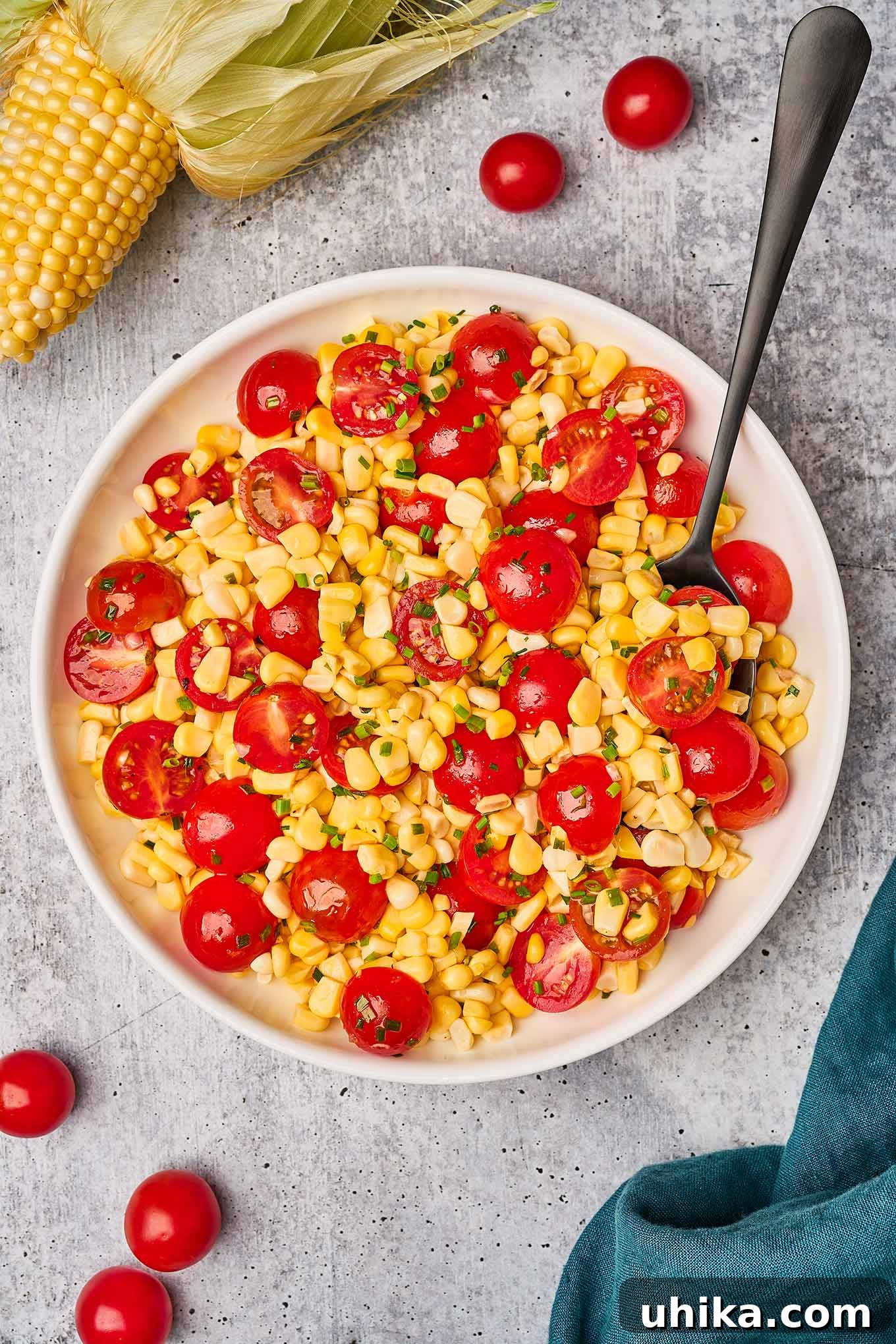 Fresh corn and cherry tomato salad in a serving bowl with a serving spoon, surrounded by a linen napkin, corn with the husk on and cherry tomatoes.