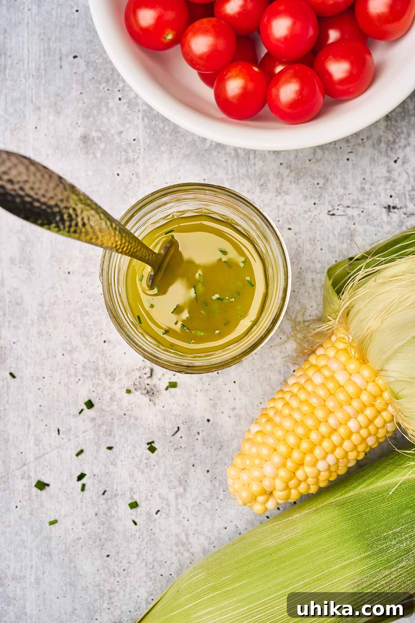 Fresh corn on the cob in the husks, cherry tomatoes, and a jar of lemon, shallot, and chive vinaigrette on a gray cement background.