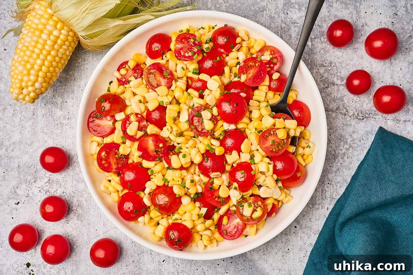 An overhead view of corn and tomato salad in a serving dish, surrounded by extra salad ingredients on a gray cement background.