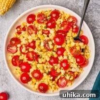 Fresh corn and cherry tomato salad in a serving bowl with a serving spoon, surrounded by a linen napkin, corn with the husk on and cherry tomatoes.