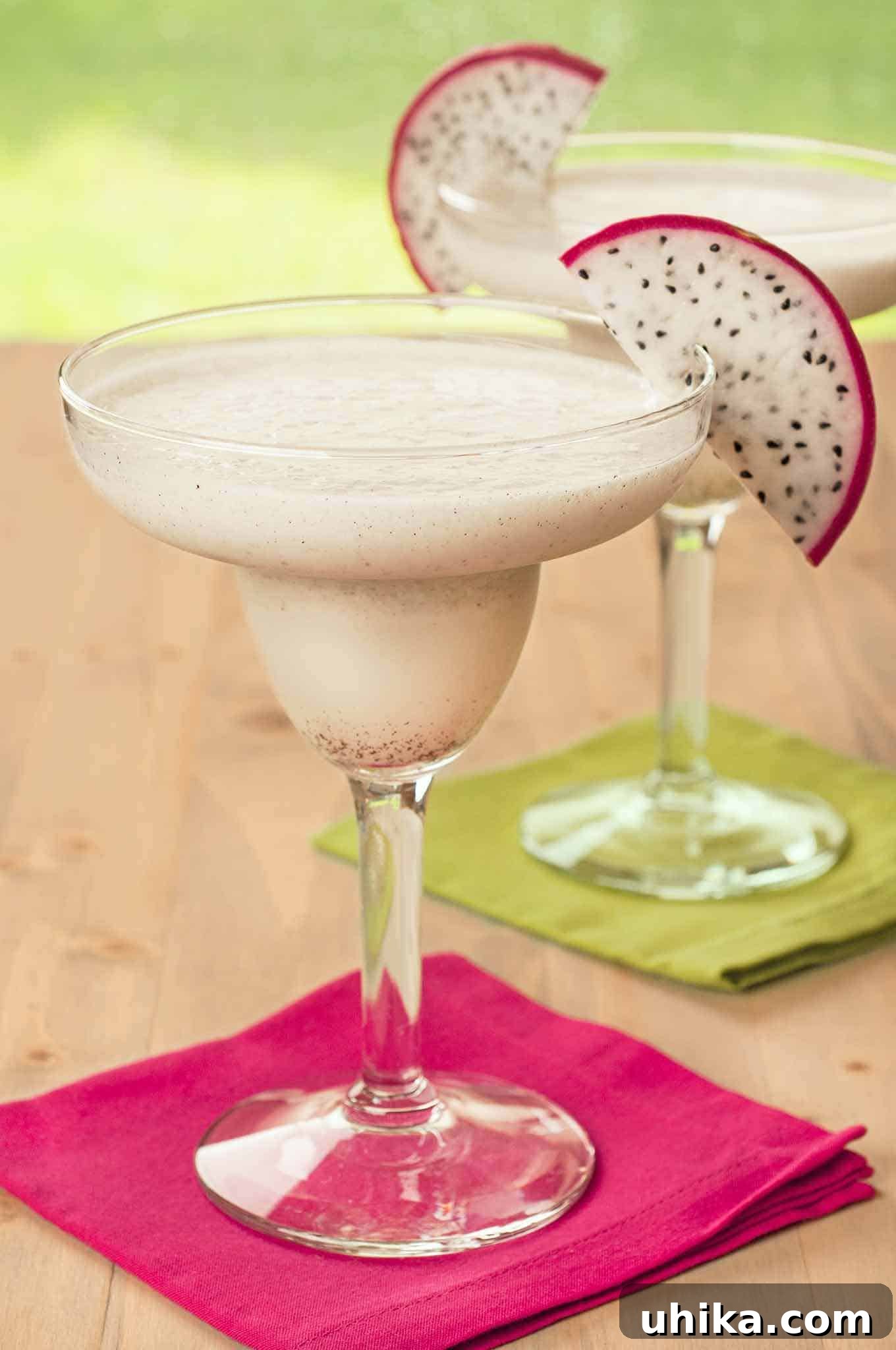 Pitaya Paradise Margarita 2 Two pristine white, frozen dragon fruit margaritas in elegant clear margarita glasses, each adorned with a vibrant slice of dragon fruit. They are placed on brightly colored pink and green cocktail napkins, highlighting their refreshing tropical appeal.