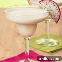 Two white, frozen margaritas made with dragon fruit in a clear margarita glass, each garnished with a slice of dragon fruit on brightly colored pink and green cocktail napkins.