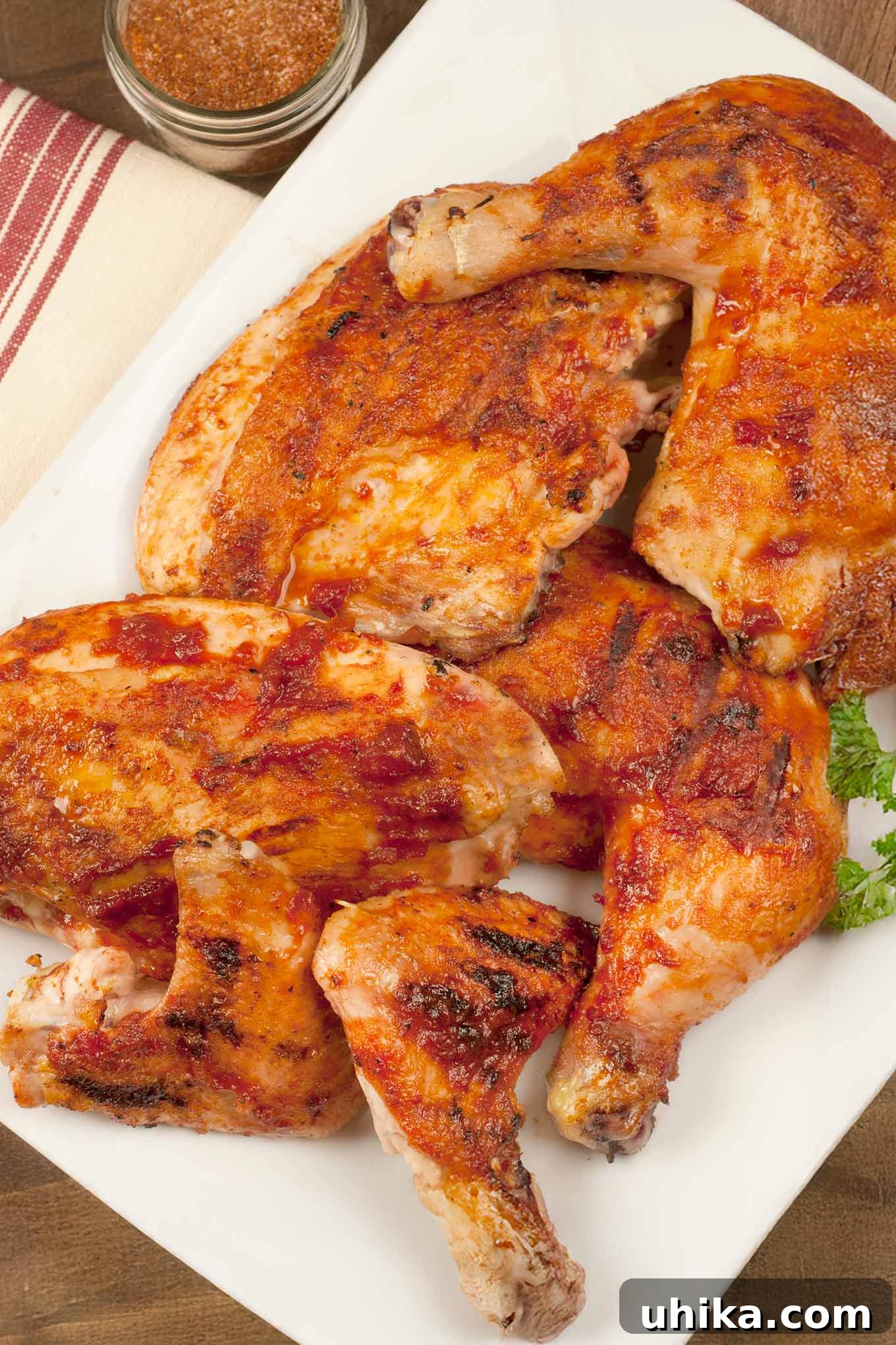 Perfectly Grilled, Ultra Juicy BBQ Chicken 2 A beautifully arranged white serving platter filled with various parts of perfectly grilled BBQ chicken, including succulent leg quarters, tender breasts, and crispy wings. A fresh sprig of parsley garnishes the side, and a jar of homemade BBQ spice rub is subtly blurred in the background, hinting at the key to its rich, smoky-sweet flavor. This image perfectly captures the essence of a delicious, home-cooked barbecue feast, ready to be enjoyed.