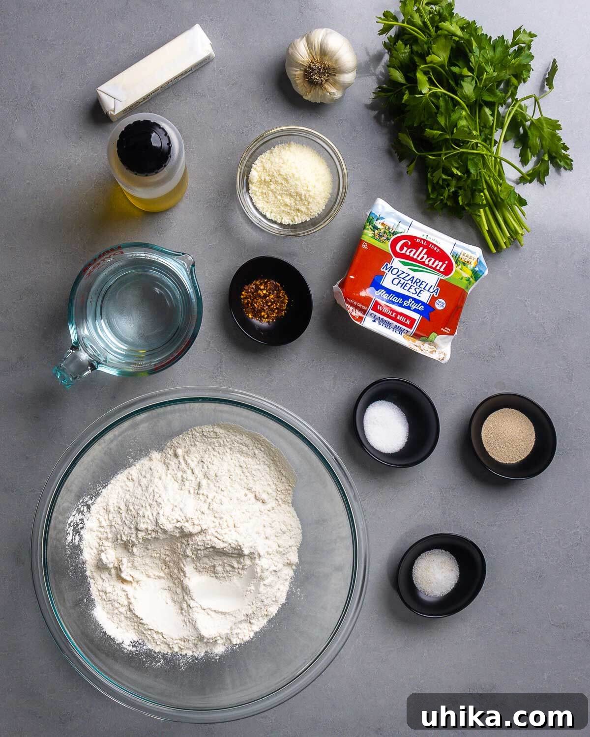 Gooey Garlic Stuffed Cheese Rolls 4 A clear overhead shot displaying all necessary ingredients for cheesy garlic rolls: various flours, yeast, salt, sugar, olive oil, a stick of butter, fresh garlic, parsley, red pepper flakes, grated Pecorino Romano, and shredded mozzarella cheese.