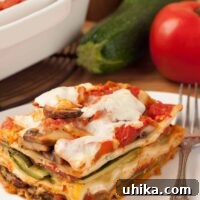 A serving of vegetable lasagna, layered zucchini, eggplant, mushrooms, and cheese, on a plate with a fork, a zucchini, tomato, and casserole of lasagna in the background.