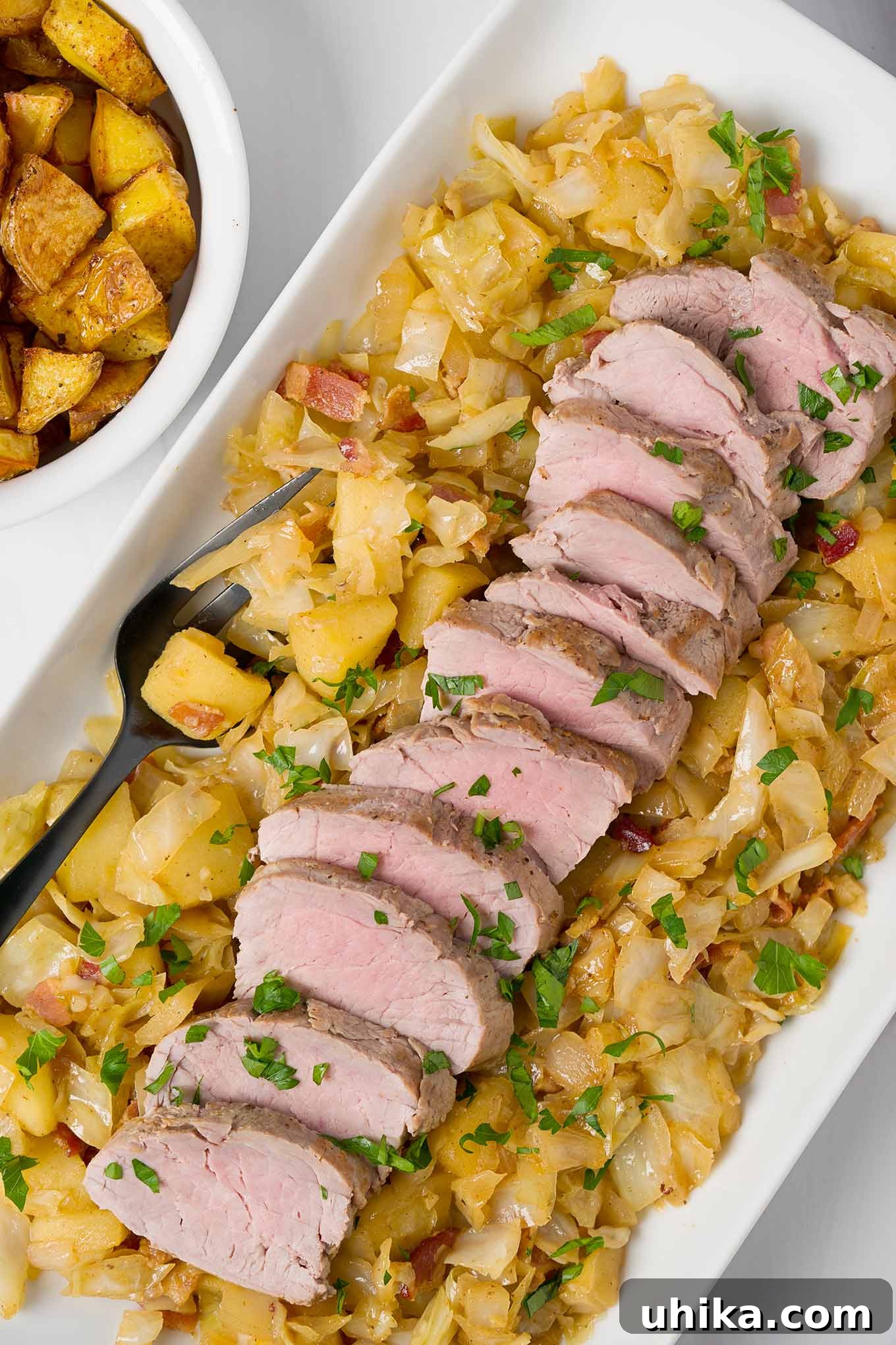 A platter of braised pork tenderloin, sliced and served on a bed of cabbage, apples, and bacon, a bowl of roasted potatoes on the side.