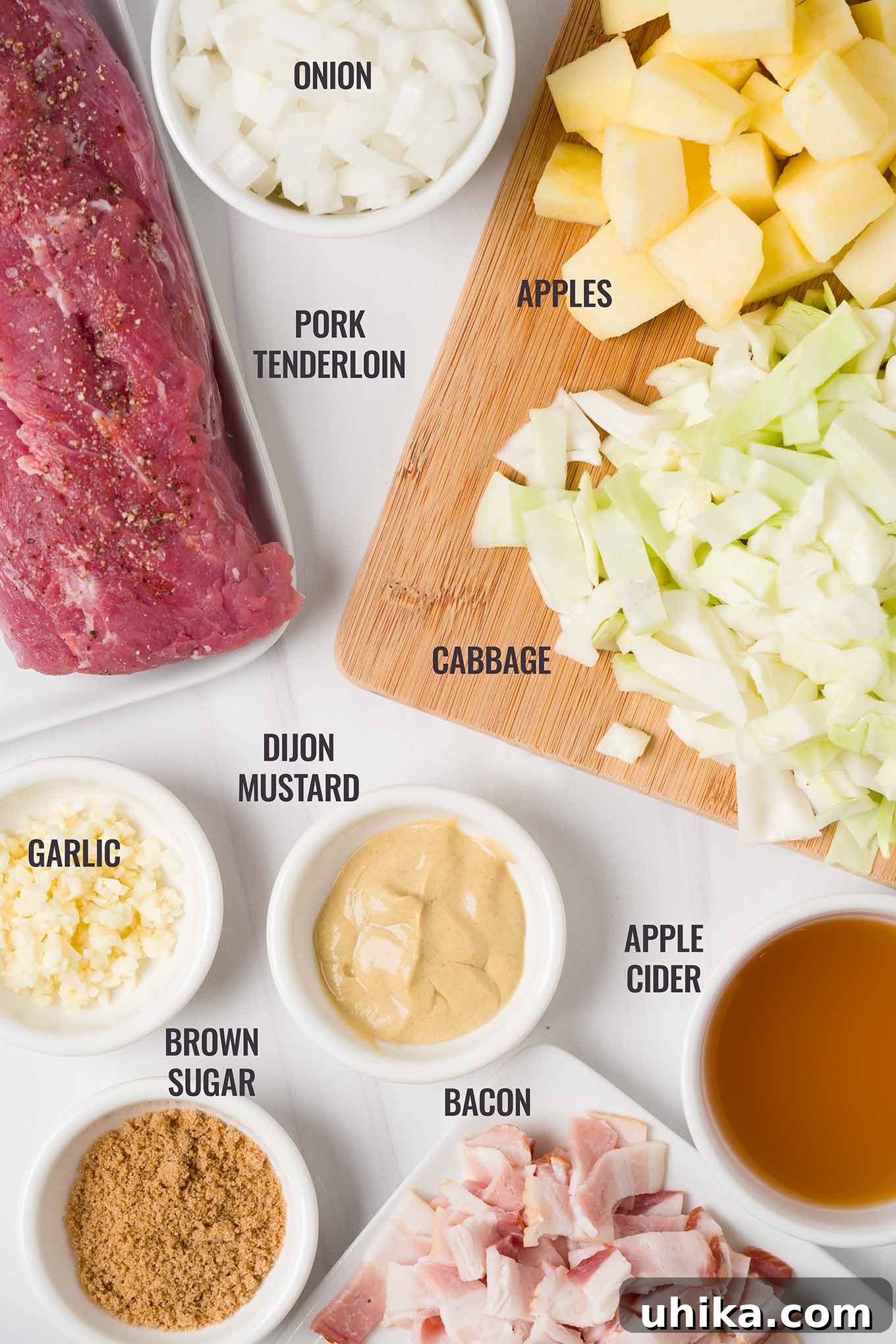 A labeled, overhead view of the ingredients for braised pork tenderloin including apples, cabbage, bacon, brown sugar, Dijon mustard, and apple cider.